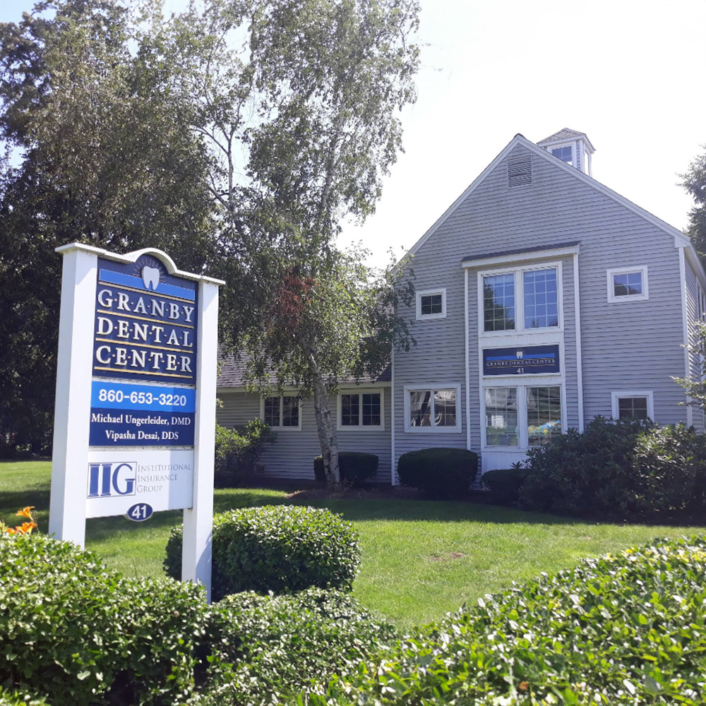 The image depicts a two-story dental practice building with a sign in front reading Granby Dental Center and a real estate sign indicating the property is for sale, set against a clear sky on a sunny day.