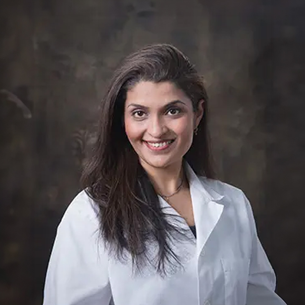 A woman wearing a white lab coat stands against a dark background with a focused expression.