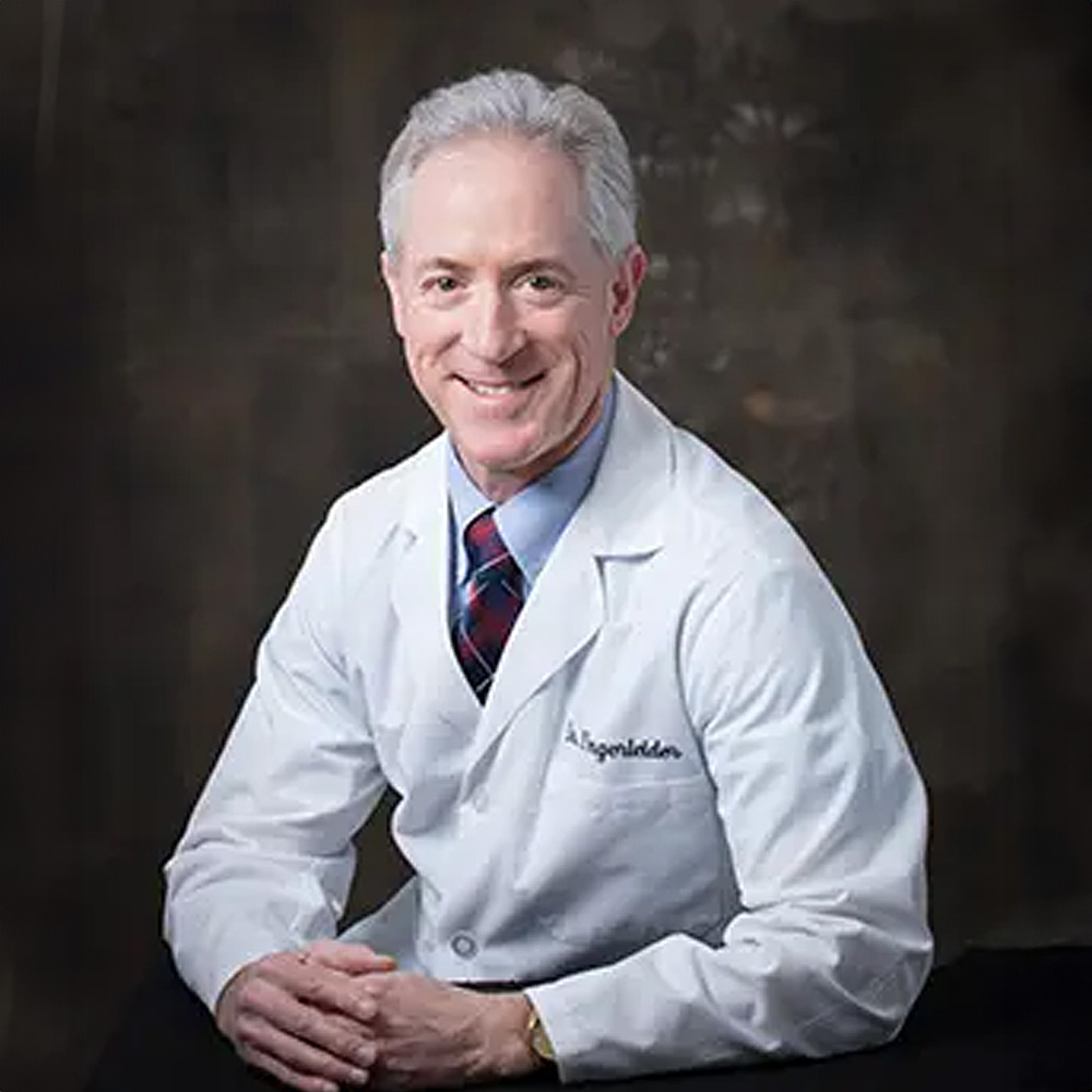 The image shows a man posing for a portrait he is wearing a white lab coat with name tags and appears to be a professional, possibly a scientist or academic, standing against a dark background with his arms crossed.