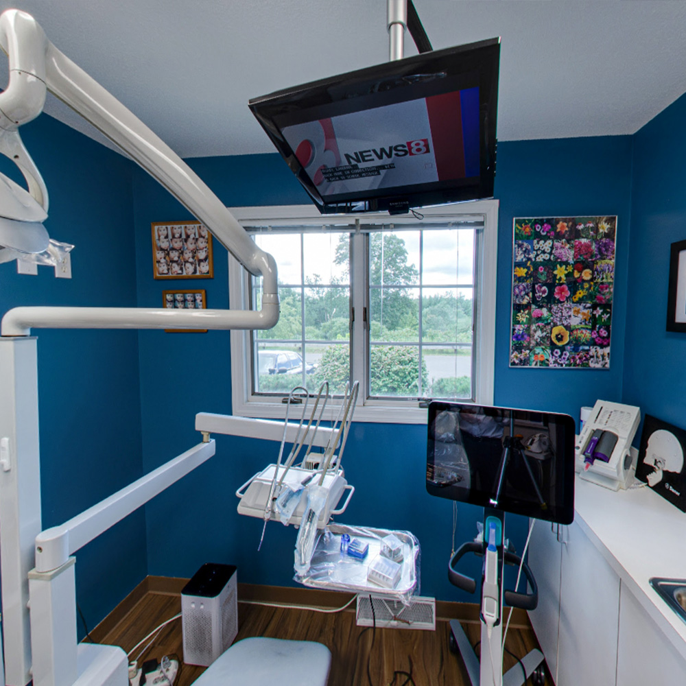 The image depicts an interior view of a dental office with modern equipment, including a treatment chair, dental station, and patient monitor.